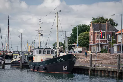 Terschelling Hafen: Ein schwarzes Boot am Anleger neben historischen Gebäuden, mit grünen Bäumen und Wolken im Hintergrund.