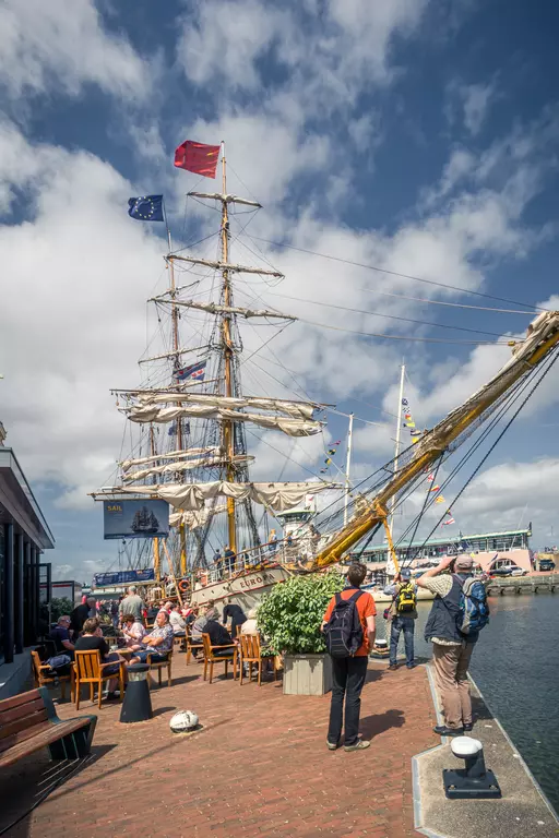 Sailing ship in Harlingen: A large ship adorned with flags is docked, surrounded by people and outdoor dining areas.