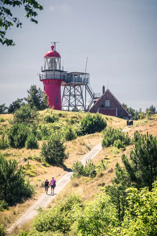 Lighthouse on Vlieland: Two people walk along a path, surrounded by hills and trees, with a red lighthouse in the background.