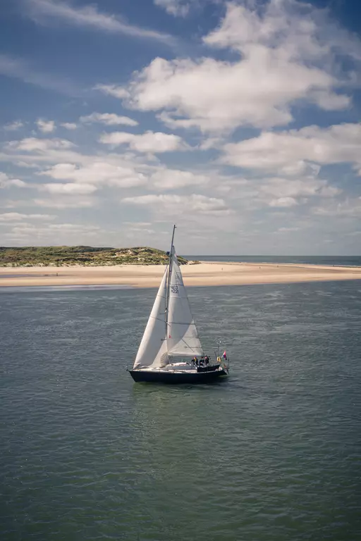 A sailboat with white sails glides through calm waters, with a sandy beach and blue sky in the background.