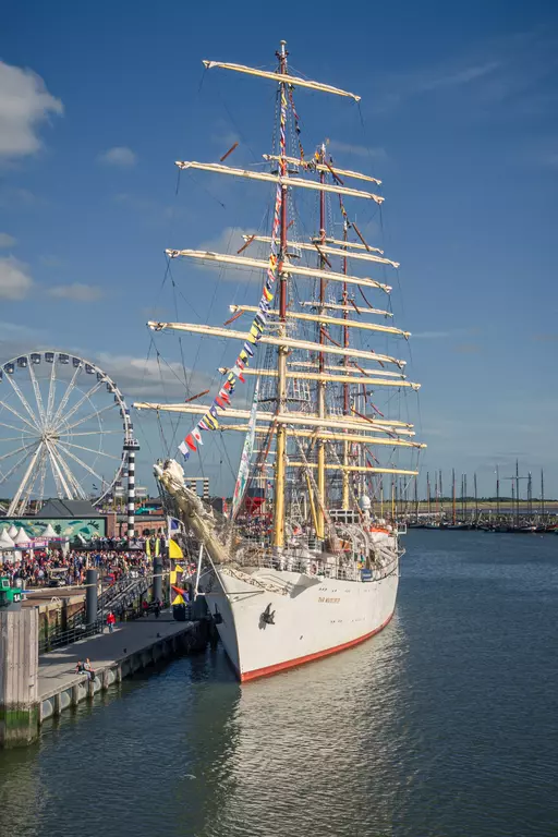 A large multi-masted sailing ship is docked at the harbor, surrounded by crowds, with a Ferris wheel in the background.