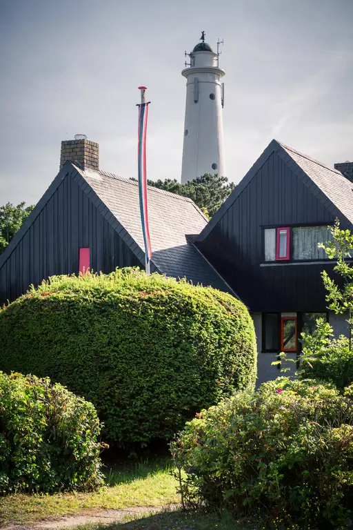 Ein modernes, schwarzes Haus mit roten Fenstern, umgeben von grünen Büschen, im Hintergrund ein hoher weißer Leuchtturm.