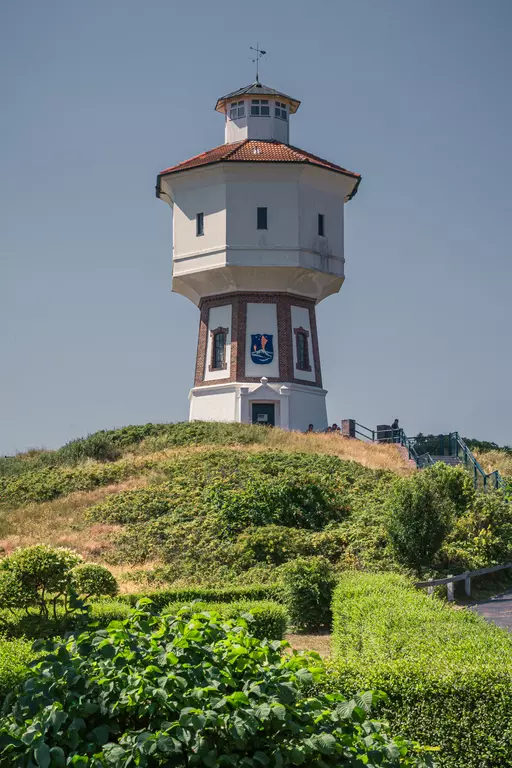 Ein historischer Wasserturm steht auf einem Hügel, umgeben von üppigem Grün und Sträuchern, unter klarem Himmel.