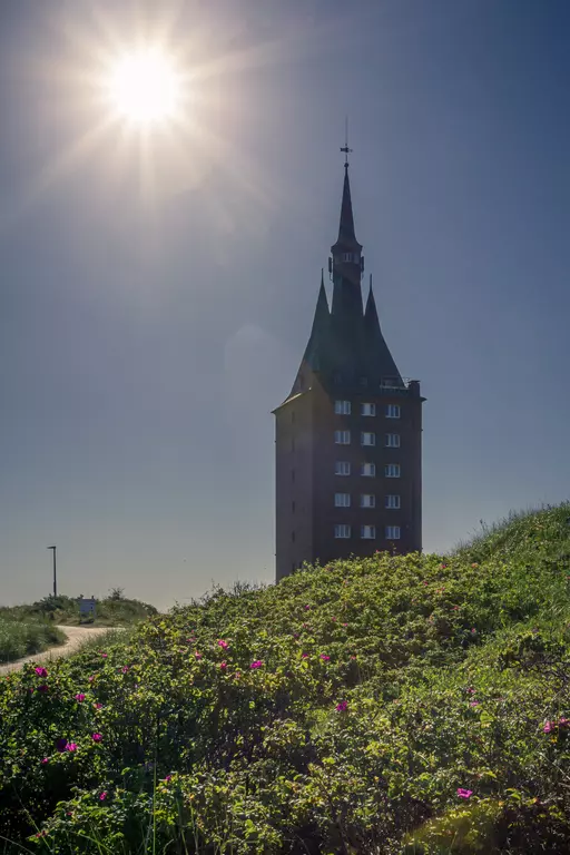 Ein hoher Turm mit Spitzdach steht auf einer Hügelkuppe, umgeben von blühendem Grün und strahlendem Sonnenlicht.