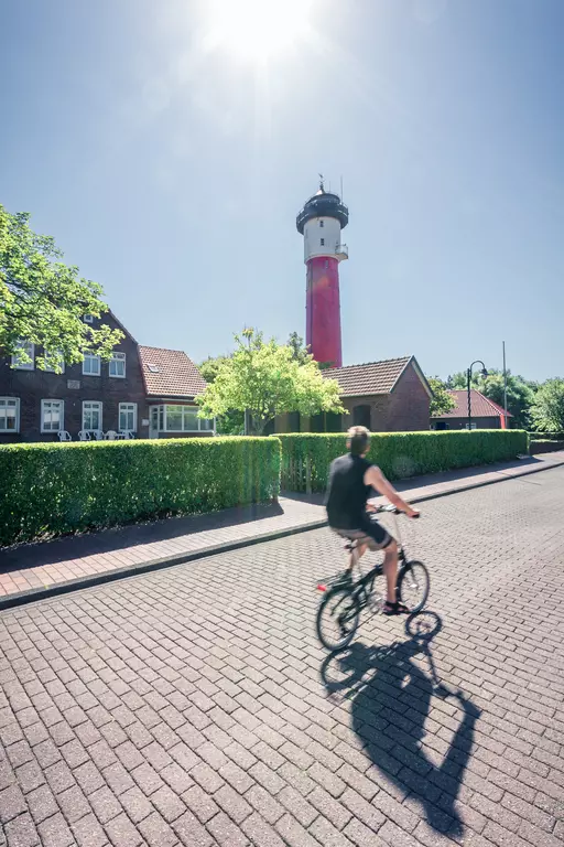 Radfahrer auf einem gepflasterten Weg, vorbei an einem roten Leuchtturm und umgeben von grünen Bäumen und Häusern.