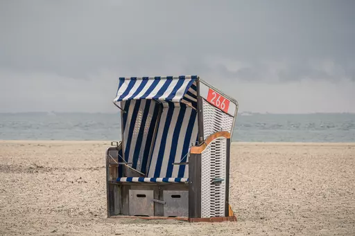 Ein Strandkorb mit blauen und weißen Streifen steht einsam am Strand, umgeben von feinem Sand und Blick auf das Wasser.