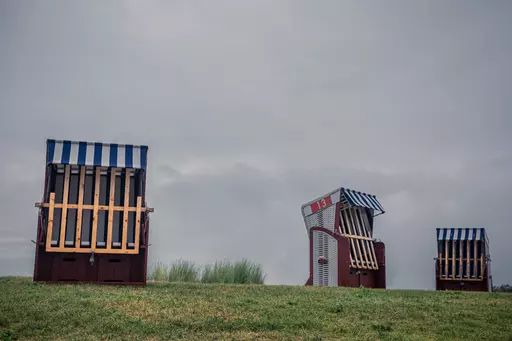 Drei Strandkörbe mit blauen und weißen Streifen stehen auf einem Hügel unter einem bewölkten Himmel. Grüne Wiese im Vordergrund.