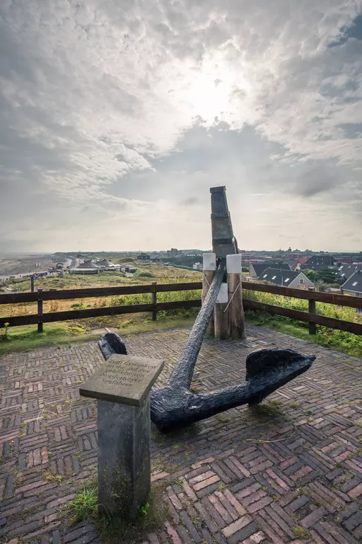 Eine große, schwarze Ankerkonstruktion steht auf einem gepflasterten Platz, umgeben von einer Holzgeländeranlage und Blick auf die Landschaft.