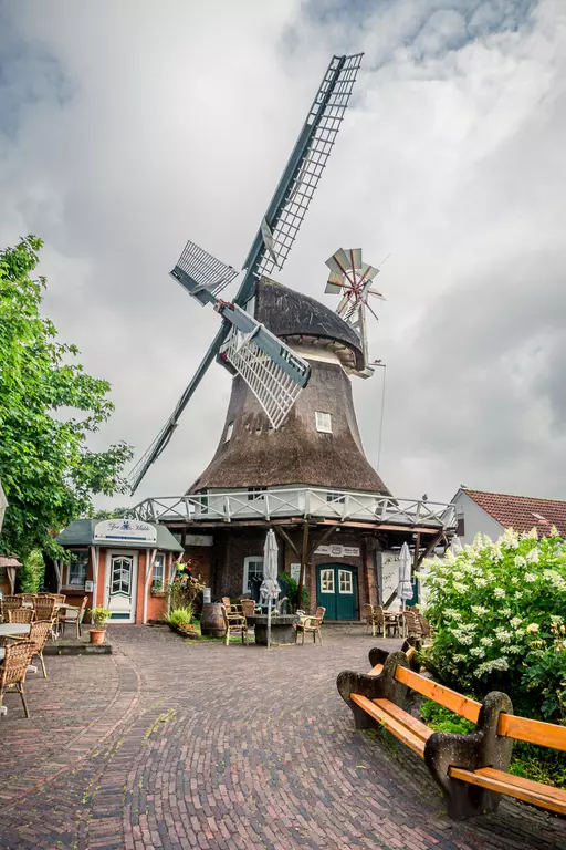 Ein großer, historischer Windmühlenbau mit einem Strohdach, umgeben von Bäumen und einer gepflasterten Terrasse mit Tischen und Bänken.