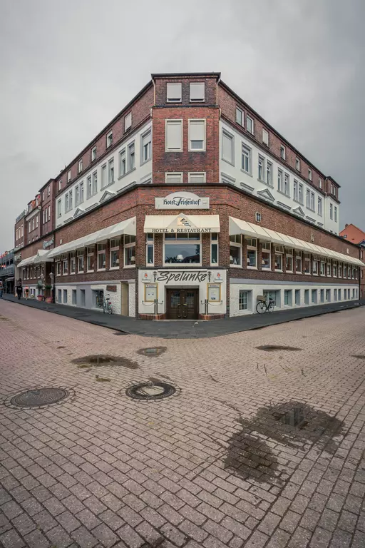 Ein dreiseitiges, historisches Gebäude mit einem Restaurant und mehreren Fenstern. Es ist bewölkt und der Boden ist nass.