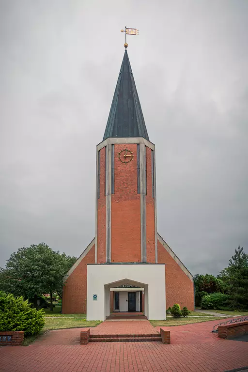 Ein rot-brauner Kirchturm mit einem spitzen Dach und einer Wetterfahne, umgeben von Bäumen und einem gepflasterten Weg.