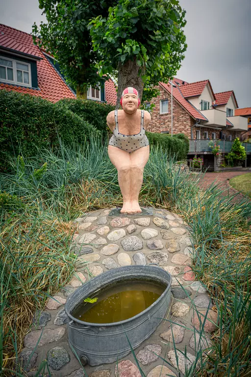 Eine Figur in Schwimmbekleidung steht neben einem steinigen Weg und einem zinnernen Behälter, umgeben von Gras und Häusern.