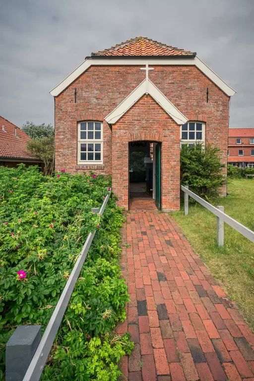 A red brick building with a gabled roof, surrounded by a paved path and green shrubs leading to the entrance door.