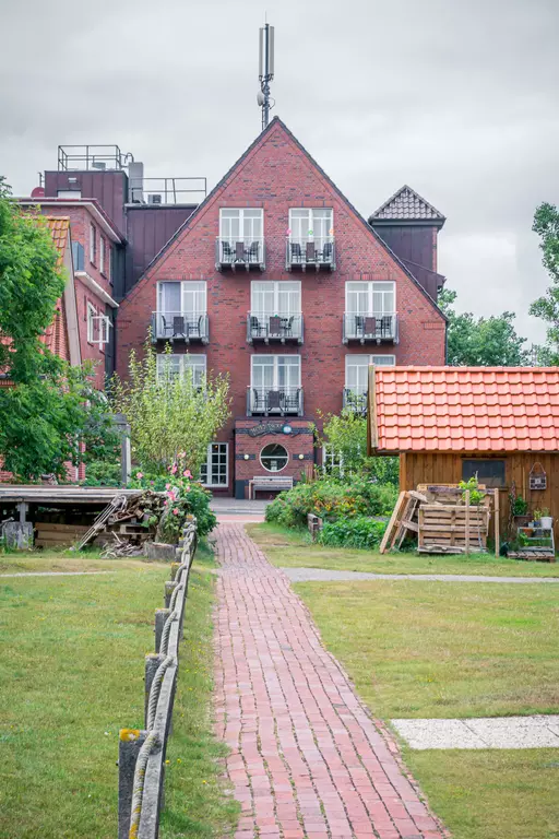 A brick walkway leads to a red brick building with balconies. Trees and a wooden structure are in the foreground.