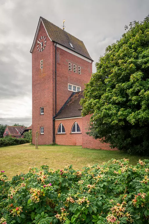 A red brick tower with a clock face is surrounded by shrubs and trees under a cloudy sky.