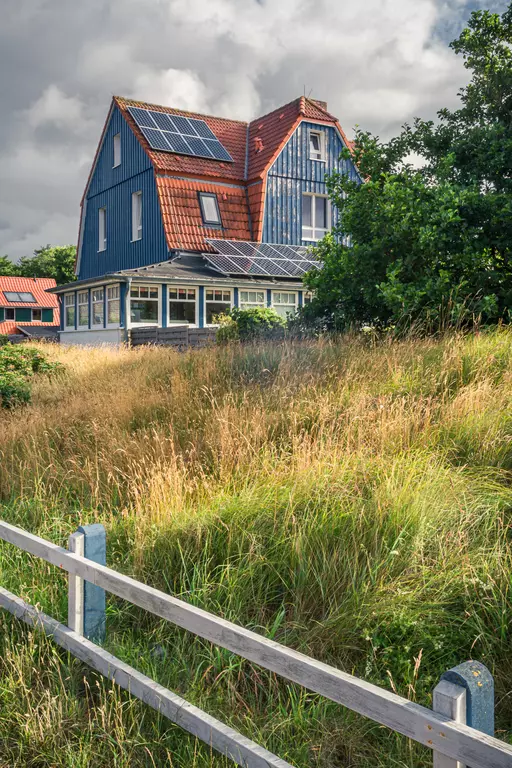 The image depicts a blue house with a red roof, surrounded by green grass and bushes under a cloudy sky.