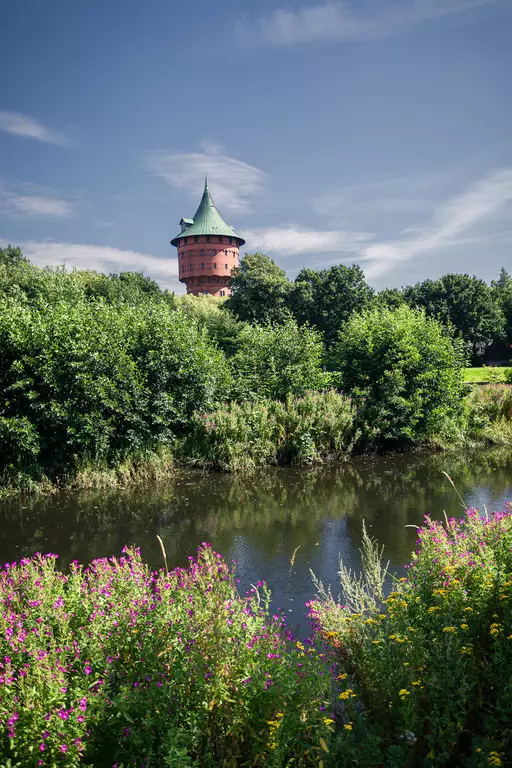 Blick auf einen historischen Turm inmitten von Bäumen und bunten Blumen am Ufer eines ruhigen Gewässers.