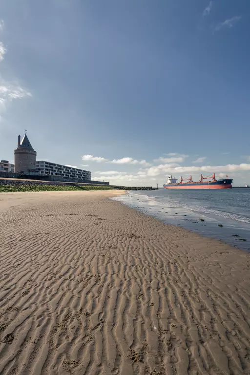 A gentle sandy beach with ripples, a large ship in the background, and a historic tower building. Clouds in a blue sky.