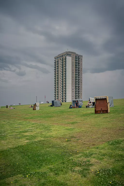 A green lawn with several beach chairs in the foreground and a tall residential building in the background under a cloudy sky.