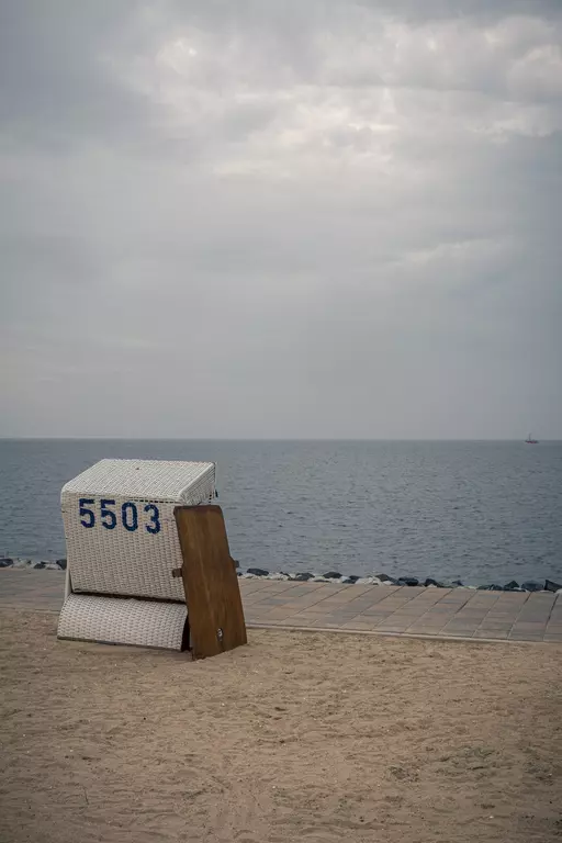 A beach chair numbered 5503 sits on the sand, overlooking calm water with a gray sky in the background.