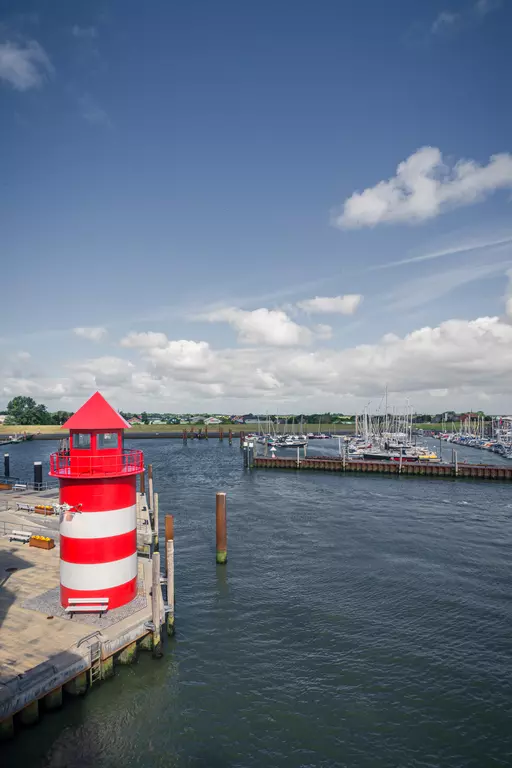 Leuchtturm auf Föhr: Ein roter und weiß gestreifter Leuchtturm steht an einer Hafenmole, umgeben von Booten und ruhigem Wasser.