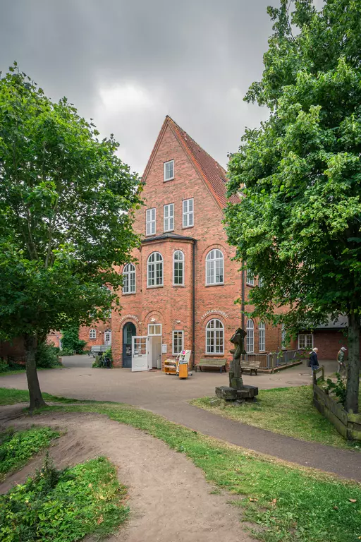 Föhr: Rote Backsteinarchitektur mit großen Fenstern und umgebenden Bäumen, Weg führt zum Eingang.