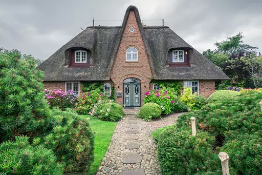 Strandhaus auf Sylt: Ein charmantes, reetgedecktes Haus umgeben von bunten Blumen und grünen Sträuchern, mit einem gepflasterten Weg.