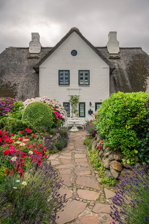 Strandhaus auf Sylt: Ein weißes Reetdachhaus mit blau gerahmten Fenstern, umgeben von bunten Blumen und einem gepflasterten Weg.