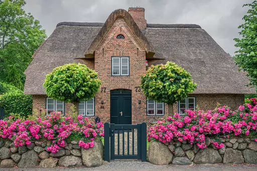 Haus in Sylt: Ein traditionelles Reetdachhaus mit Blumenbeeten und Gehweg aus Steinen. Grüne Bäume flankieren den Eingang.