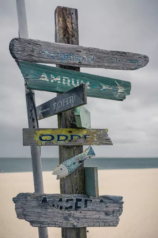 Strand Beschilderung auf Sylt: Ein Holzschild mit verschiedenen Richtungspfeilen zu benachbarten Inseln, verziert mit einer Fischzeichnung.