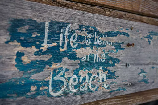 Wettergegerbtes Holzschild mit dem Text Life is better at the Beach in blauer und weißer Farbe, teils abgerieben und verwittert.