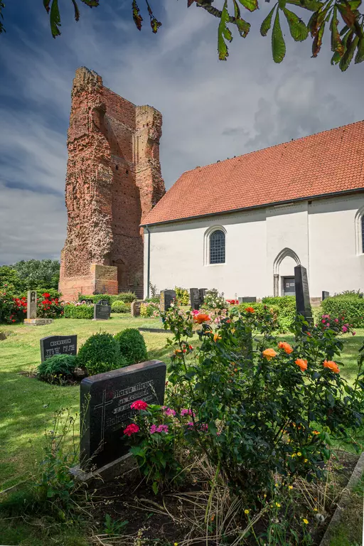 Blick auf eine alte Ruine neben einer weißen Kirche, umgeben von Blumen und Gräbern im Gartenbereich.