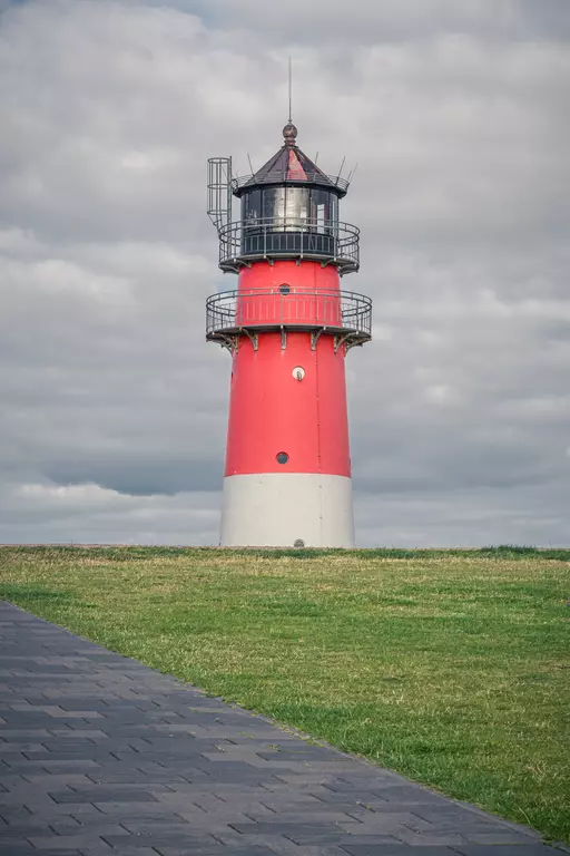 A red lighthouse with a black top stands on a grassy area under a cloudy sky. A pathway leads up to the tower.