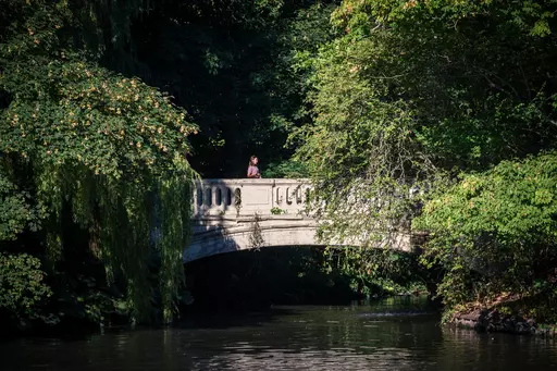 A stone bridge over a calm stream, surrounded by green trees, with a person standing on the bridge.