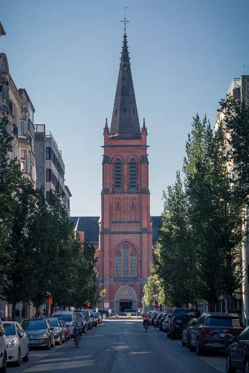 A tall, red brick church with a pointed steeple stands at the end of a tree-lined street, with cars parked along the side.