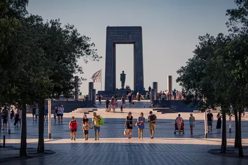 Several people walk across a wide plaza surrounded by trees, with a monumental arch and statue in the background.