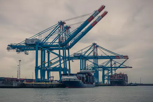 Große blaue Kräne heben Container in einem Hafen; zwei Frachtschiffe liegen am Kai, der Himmel ist bewölkt.