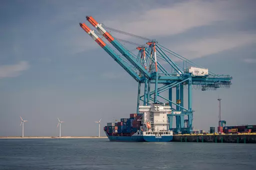 Ein großes Containerschiff liegt im Hafen, während Kräne Container entladen. Windräder sind im Hintergrund sichtbar.