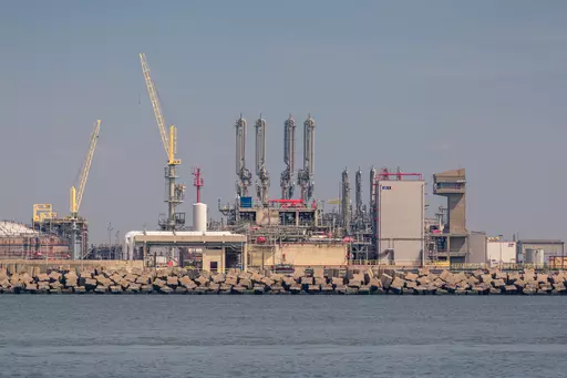 Industrielle Anlage mit hohen Türmen, Rohren und Kränen, die am Ufer stehen, umgeben von Wasser und Felsen.