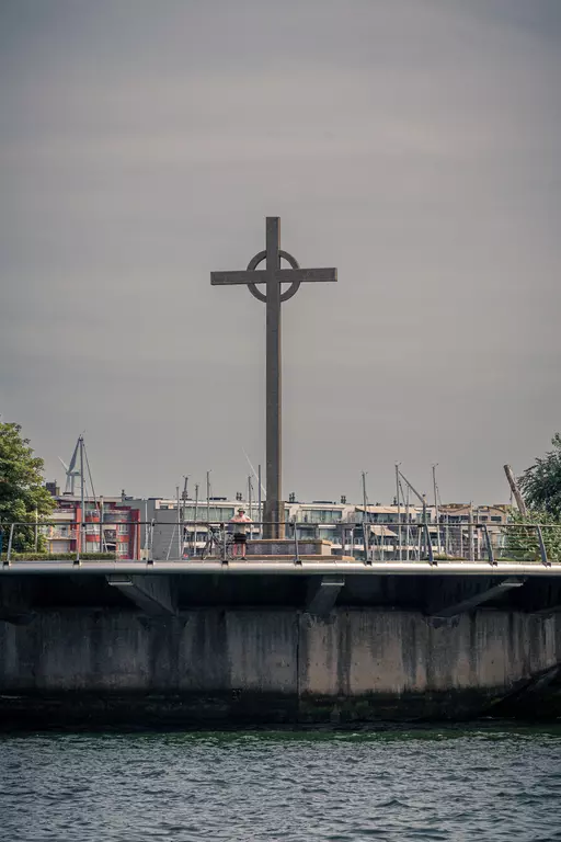 Ein großes Kreuz steht auf einem stabilen Fundament am Wasser, umgeben von Bäumen und Gebäuden im Hintergrund.