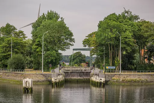 A bridge over a waterway, surrounded by lush greenery and wind turbines in the background, reflecting in the water.