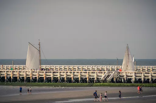 Two sailboats are sailing on the water while people walk on a wooden pier. The sky is cloudy.