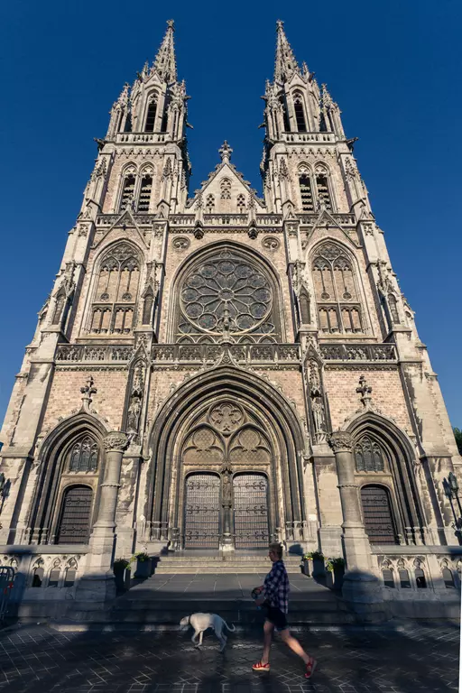 A large Gothic cathedral with two tall towers and a large rose window. A person walks by with a dog.