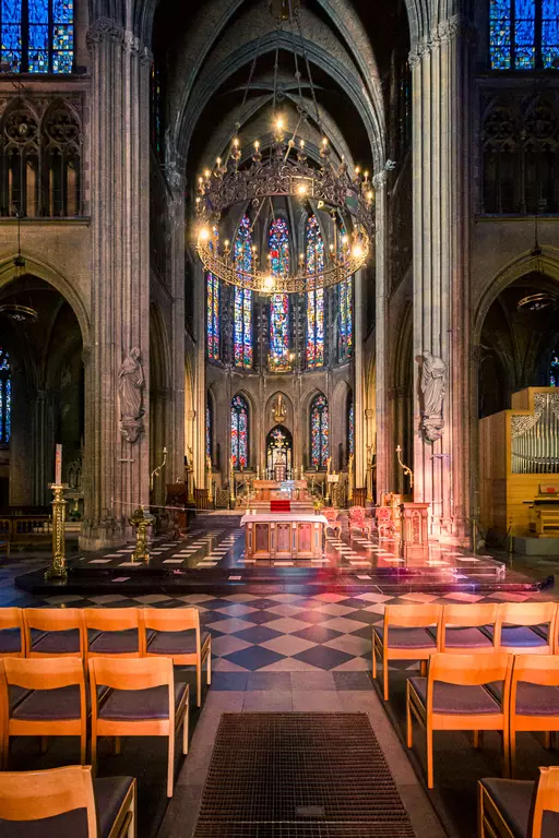 Bright church interior featuring stained glass windows, a central altar area, and wooden chairs in the foreground.