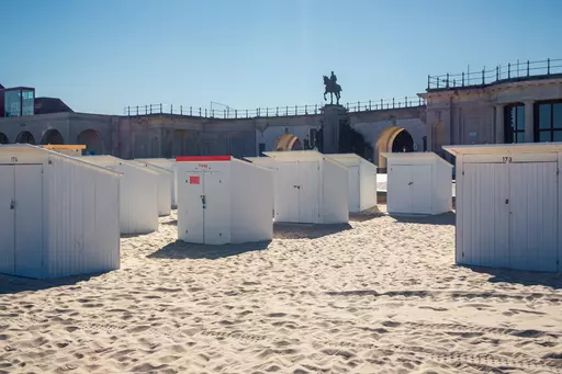 Rows of white beach huts on sandy ground, with a historic building and a statue on a pedestal in the background.