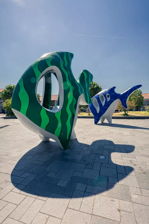 A large, colorful fish sculpture in green and blue hues stands on a paved surface, surrounded by trees.