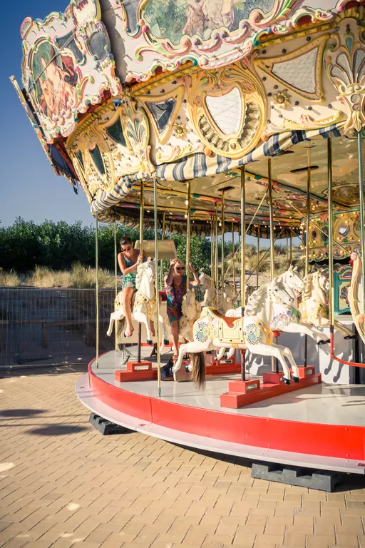 Two children ride a colorful carousel with white horses, surrounded by bright sunshine and greenery in the background.