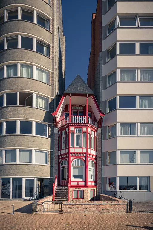 A colorful three-story house in red and white stands between two modern apartment buildings.