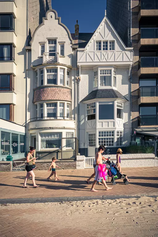 Several people walk along a sandy path between a historic and a modern building, some in swimwear.