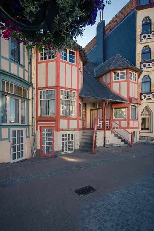 Colorful, historic buildings with red and green accents line a quiet street. Stairs lead up to an entrance area.